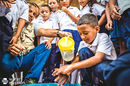 photo of a child washing their hands as adult pours pitcher of water on his hands
